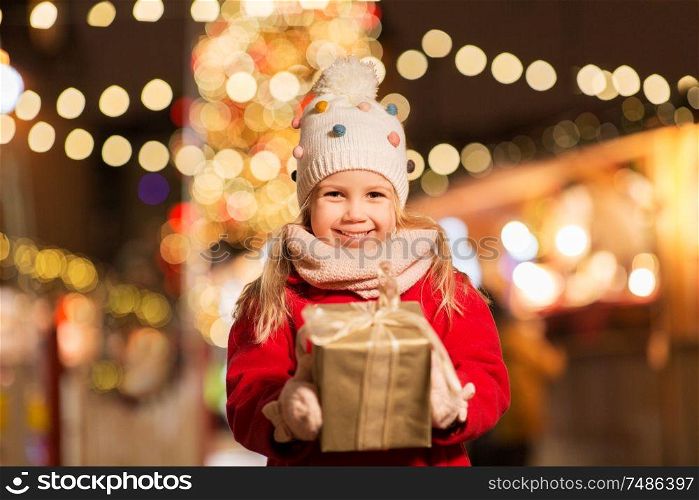 holidays, childhood and people concept - happy little girl with gift box at christmas market in winter evening. happy girl with gift box at christmas market
