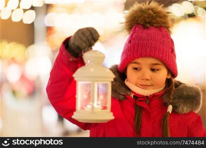 holidays, childhood and people concept - happy little girl with lantern at christmas market in winter evening. happy little girl at christmas with lantern market