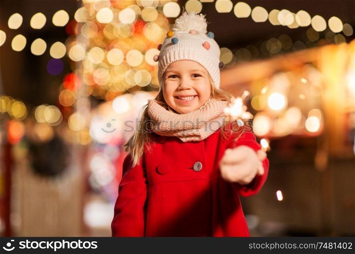holidays, childhood and people concept - happy little girl with sparkler at christmas market in winter evening. happy girl with sparkler at christmas market