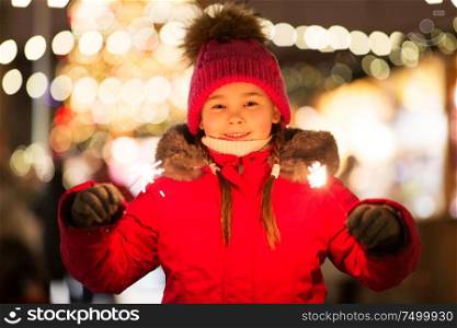 holidays, childhood and people concept - happy little girl with sparklers at christmas market in winter evening. happy girl with sparklers at christmas market
