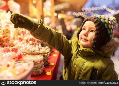 holidays, childhood and people concept - little boy choosing sweets at christmas market candy shop in winter evening. little boy at christmas market candy shop