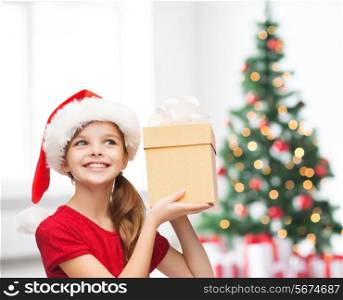 holidays, childhood and people concept - smiling girl in santa helper hat with gift box over living room and christmas tree lights background