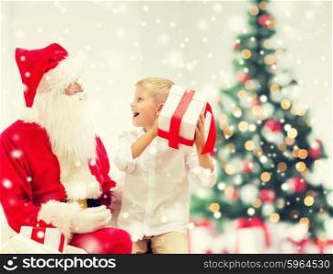 holidays, childhood and people concept - smiling little boy, santa claus with gifts over living room, christmas tree and snow background
