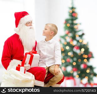 holidays, childhood and people concept - smiling little boy with santa claus and gifts over christmas tree lights background