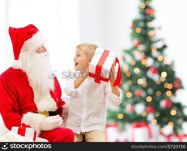 holidays, childhood and people concept - smiling little boy with santa claus and gifts over christmas tree lights background
