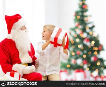 holidays, childhood and people concept - smiling little boy with santa claus and gifts over christmas tree lights background