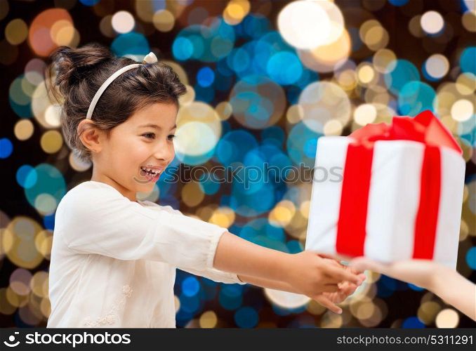 holidays, childhood and people concept - smiling little girl giving or receiving present over lights background. smiling little girl giving or receiving present