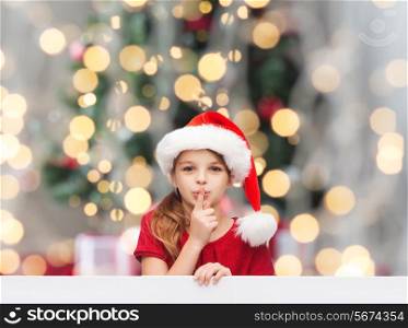 holidays, childhood and people concept - smiling little girl in santa helper hat with finger on her lips over christmas tree lights background
