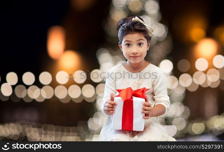 holidays, childhood and people concept - smiling little girl with gift box over christmas tree lights background. happy girl with gift box over christmas lights
