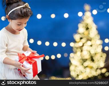 holidays, childhood and people concept - smiling little girl with gift box over christmas tree lights background. happy girl with gift box over christmas lights