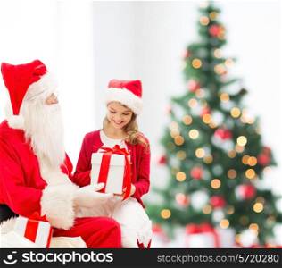 holidays, childhood and people concept - smiling little girl with santa claus and gifts over christmas tree lights lights background