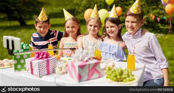 holidays, childhood and technology concept - happy kids taking picture by selfie stick on birthday party at summer garden. happy kids taking selfie on birthday party