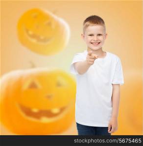 holidays, childhood, happiness, gesture and people concept - smiling little boy in white blank shirt pointing finger at you over halloween pumpkins background