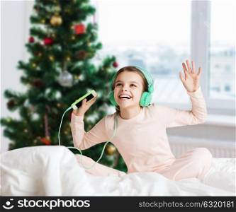 holidays, children and technology concept - happy smiling girl in headphones sitting on bed with smartphone and listening to music over christmas tree background. girl with smartphone and headphones at christmas
