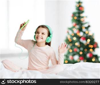 holidays, children and technology concept - happy smiling girl in headphones sitting on bed with smartphone and listening to music over christmas tree background. girl with smartphone and headphones at christmas