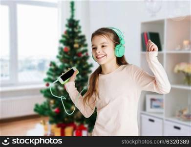 holidays, children and technology concept - happy smiling girl in headphones with smartphone and listening to music and dancing over christmas tree background. girl with smartphone and headphones at christmas
