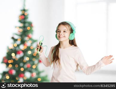 holidays, children and technology concept - happy smiling girl in headphones with smartphone and listening to music and dancing over christmas tree background. girl with smartphone and headphones at christmas