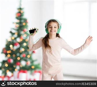 holidays, children and technology concept - happy smiling girl in headphones with smartphone and listening to music and dancing over christmas tree background. girl with smartphone and headphones at christmas