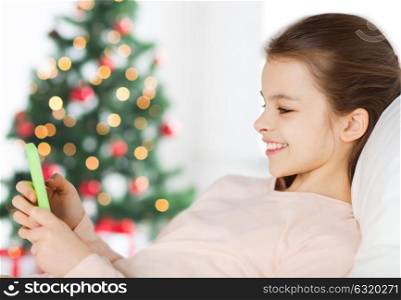 holidays, children and technology concept - happy smiling girl with smartphone lying in bed at home over christmas tree background. happy girl in bed with smartphone at christmas