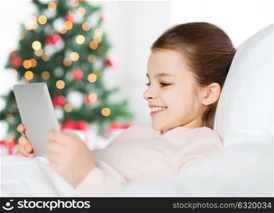 holidays, children and technology concept - happy smiling girl with tablet pc computer lying in bed over christmas tree background. happy girl in bed with tablet pc at christmas