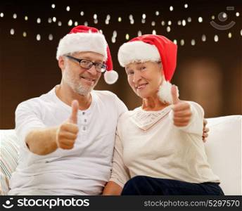 holidays, christmas, age and people concept - happy senior couple in santa hats showing thumbs up over lights background. happy senior couple in santa hats for christmas