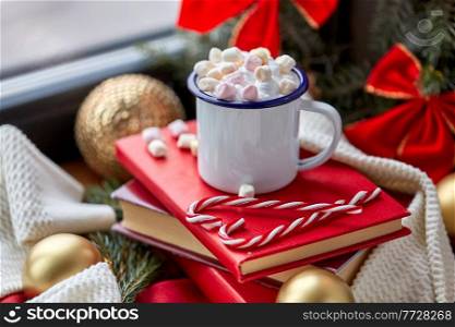 holidays, christmas and celebration concept - camp mug of whipped cream with marshmallow, candy canes, books and decorations at home. cup of whipped cream with marshmallow on christmas