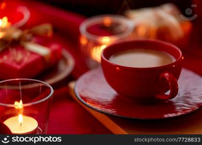 holidays, christmas and celebration concept - close up of coffee in red ceramic cup with saucer and burning candles at home. cup of coffee and candles at home on christmas