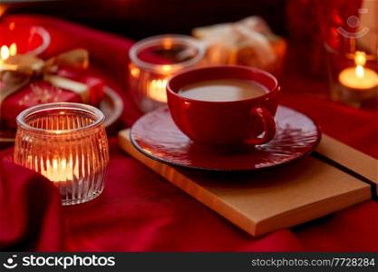 holidays, christmas and celebration concept - coffee in red ceramic cup with saucer and burning candles at home. cup of coffee and candles at home on christmas