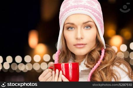 holidays, christmas and people concept - close up of young woman in winter hat with red mug over lights background. close up of woman with mug over christmas lights