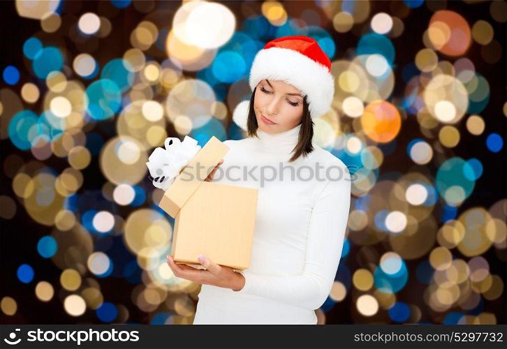 holidays, christmas and people concept - displeased woman in santa hat opening gift box over lights background. displeased woman in santa hat with christmas gift