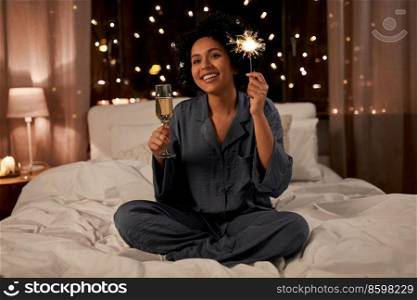 holidays, christmas and people concept - happy smiling woman in pajamas holding glass of ch&agne and extinguished sparkler sitting in bed at night. happy woman drinking ch&agne in bed at night