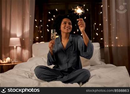 holidays, christmas and people concept - happy smiling woman in pajamas holding glass of ch&agne and extinguished sparkler sitting in bed at night. happy woman drinking ch&agne in bed at night