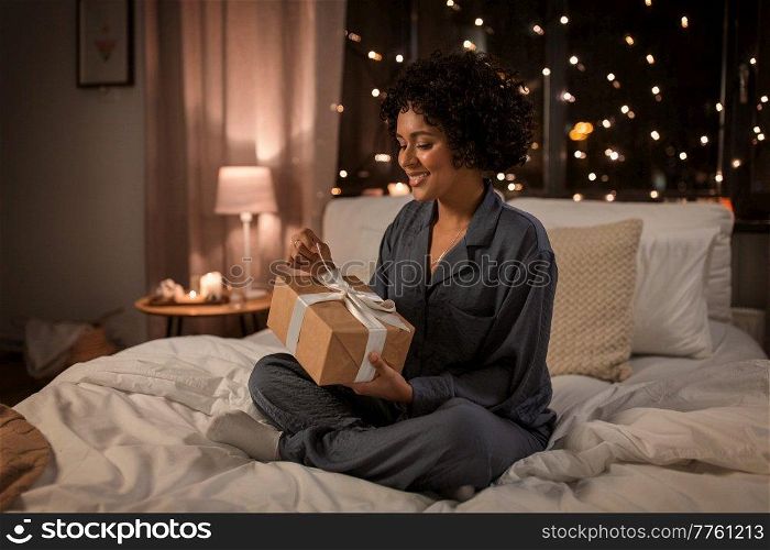 holidays, christmas and people concept - happy smiling woman in pajamas with gift box sitting in bed at night. woman in pajamas with gift sitting in bed at night