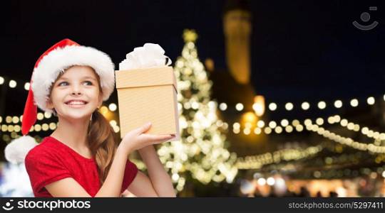 holidays, christmas and people concept - smiling little girl in santa hat with gift box over lights background. happy girl with gift over christmas tree lights