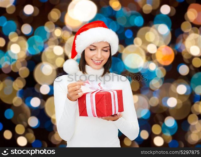 holidays, christmas and people concept - smiling woman in santa hat opening gift box over lights background. smiling woman in santa hat with christmas gift