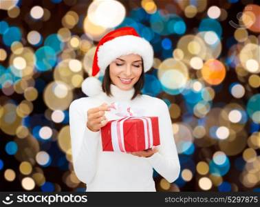 holidays, christmas and people concept - smiling woman in santa hat opening gift box over lights background. smiling woman in santa hat with christmas gift