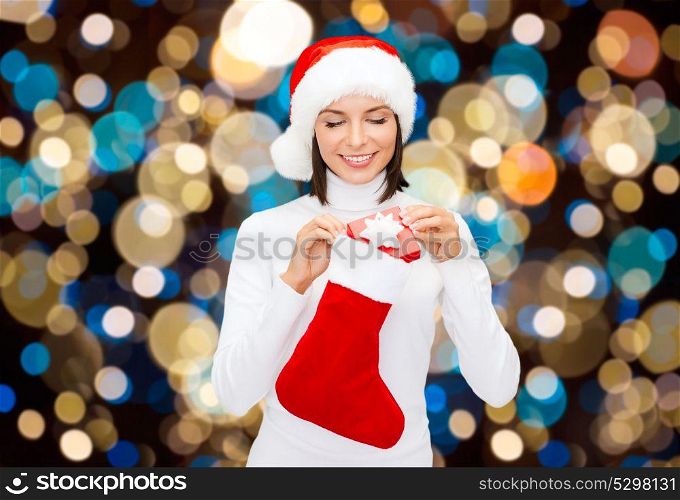 holidays, christmas and people concept - smiling woman in santa hat putting gift box into sock over lights background. woman in santa hat with christmas gift and sock