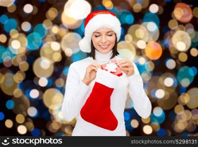 holidays, christmas and people concept - smiling woman in santa hat putting gift box into sock over lights background. woman in santa hat with christmas gift and sock