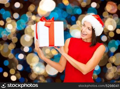 holidays, christmas and people concept - smiling woman in santa hat with gift box over lights background. smiling woman in santa hat with christmas gift