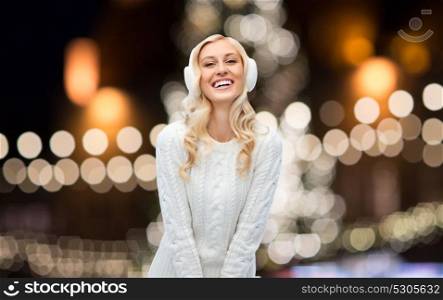holidays, christmas and people concept - smiling young woman in earmuffs and sweater over night lights background. happy woman over christmas lights