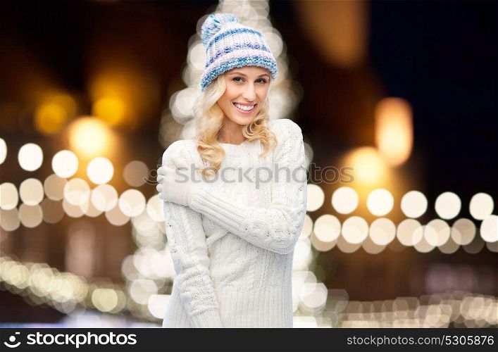 holidays, christmas and people concept - smiling young woman in winter hat, sweater and gloves over night lights background. happy woman over christmas lights