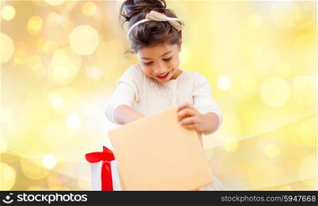 holidays, christmas, birthday childhood and people concept - smiling little girl with gift box over yellow lights background