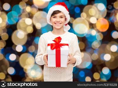 holidays, christmas, childhood and people concept - smiling happy boy in santa hat with gift box over lights background. smiling happy boy in santa hat with christmas gift