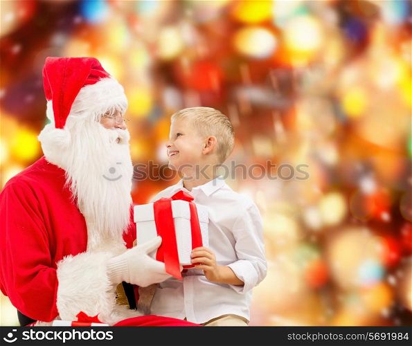 holidays, christmas, childhood and people concept - smiling little boy with santa claus and gifts over red lights background