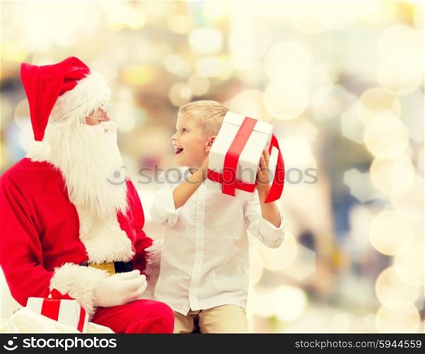 holidays, christmas, childhood and people concept - smiling little boy with santa claus and gifts over lights background