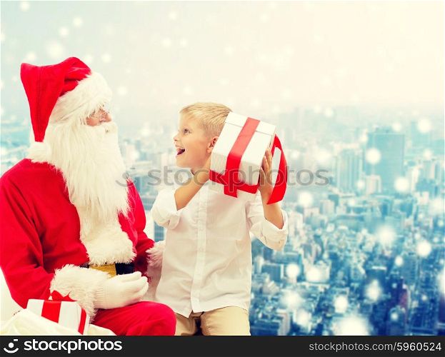 holidays, christmas, childhood and people concept - smiling little boy with santa claus and gifts over snowy city background