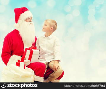 holidays, christmas, childhood and people concept - smiling little boy with santa claus and gifts over blue lights background