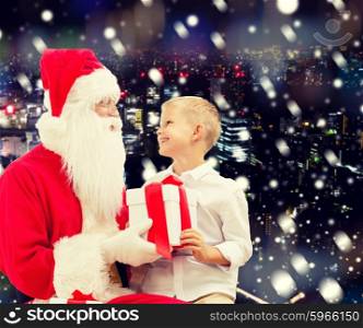 holidays, christmas, childhood and people concept - smiling little boy with santa claus and gifts over snowy city background
