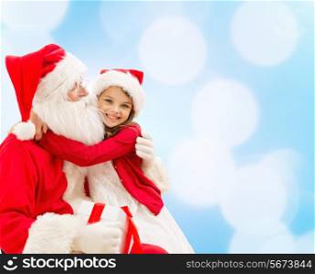 holidays, christmas, childhood and people concept - smiling little girl hugging with santa claus over blue lights background