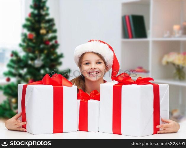 holidays, christmas, childhood and people concept - smiling little girl with gifts at home. smiling girl in santa hat with christmas gifts